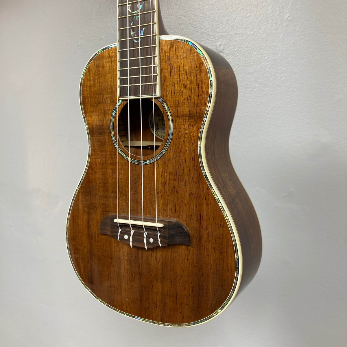 A close-up of an Oscar Schmidt OU280SWK Concert Ukulele with solid koa body, four strings, white fret markers, and abalone binding around the body and sound hole, hanging against a plain light gray wall. Includes case; used consign.