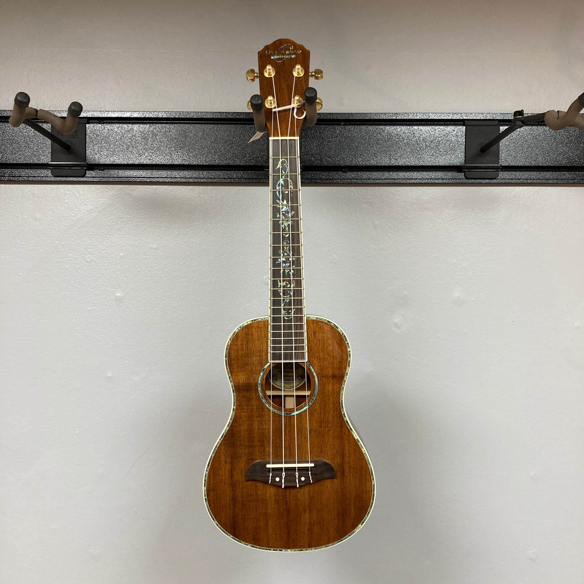 An Oscar Schmidt OU280SWK Concert Ukulele with abalone binding and decorative fretboard inlays hangs on a black wall-mounted display rack against an off-white wall.
