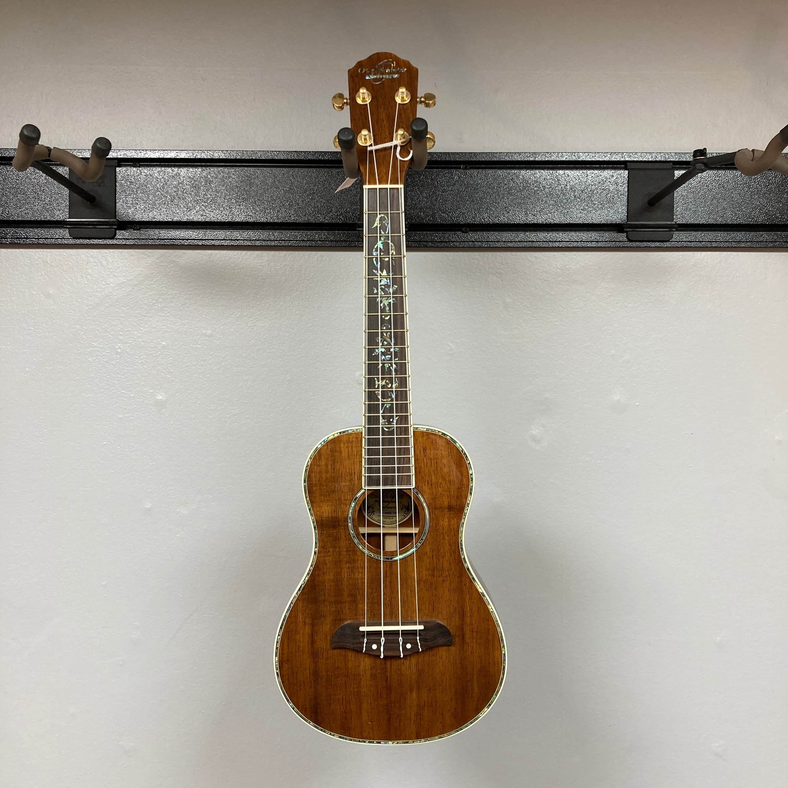 The Oscar Schmidt OU280SWK Concert Ukulele w/Case Used Consign, a brown solid koa instrument with abalone binding and decorative inlays, is shown against a plain light gray wall.