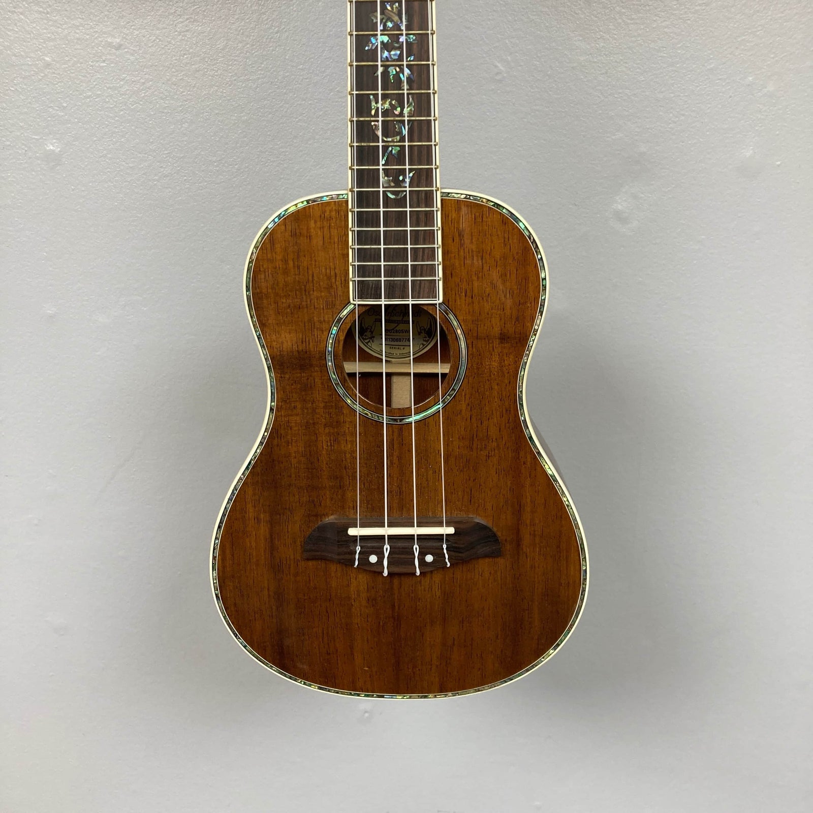 The Oscar Schmidt OU280SWK Concert Ukulele w/Case Used Consign, a brown solid koa instrument with abalone binding and decorative inlays, is shown against a plain light gray wall.