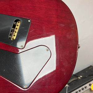 Close-up of the back of a red PRS Custom 24 Angry Larry 2011 electric guitar by PRS, featuring two black plastic control cavity covers, visible scratches, and part of an amplifier in the background.