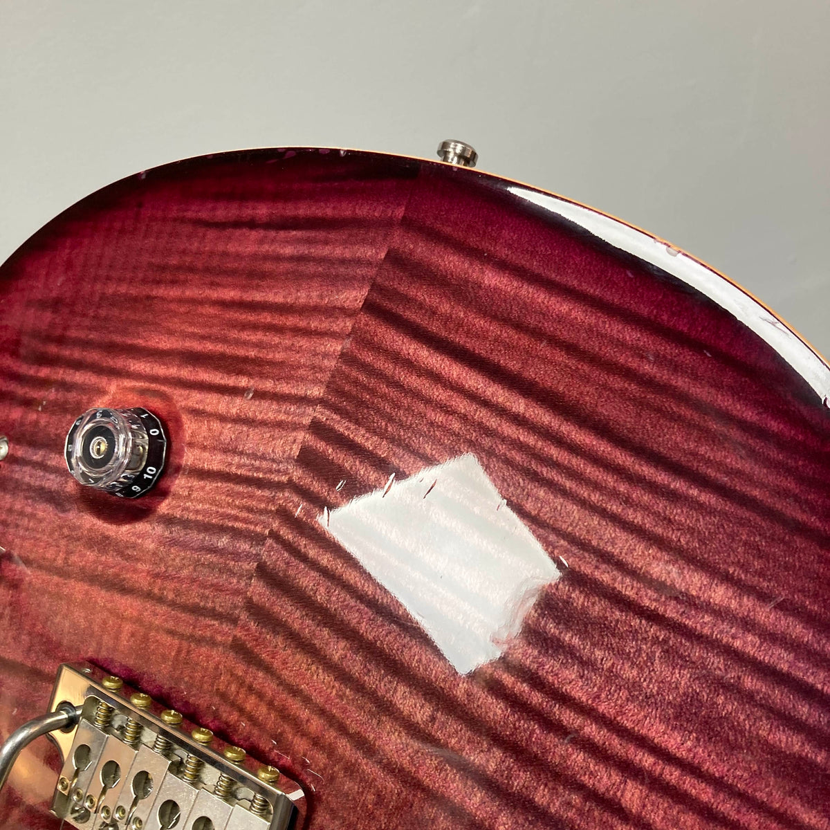 Close-up of a red PRS Custom 24 Angry Larry 2011 electric guitar body, showing a patch of damaged or missing finish near the volume knob and bridge that reveals lighter wood underneath. The area around the humbuckers is also visible, highlighting this PRS instrument’s unique character and well-played history. Includes case; used condition.