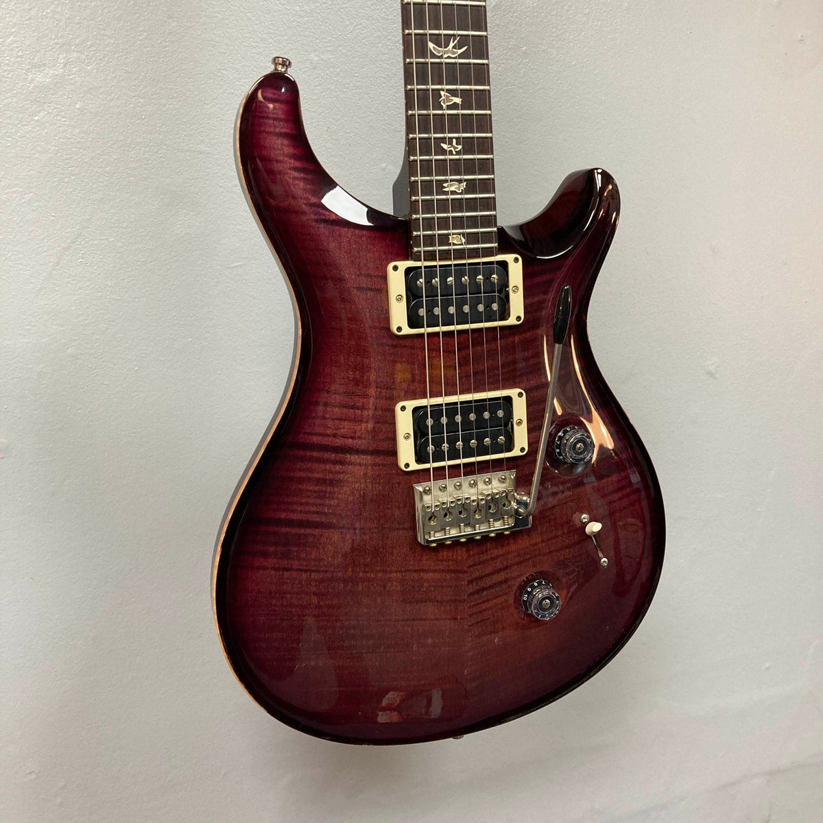 A PRS Custom 24 Angry Larry 2011 electric guitar by PRS, featuring bird inlays on the fretboard, two humbucker pickups, a tremolo bridge, and control knobs, is hanging on a light gray wall.