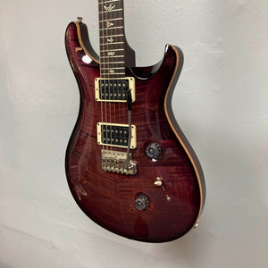 A PRS Custom 24 Angry Larry 2011 electric guitar in a rich maroon finish, featuring signature bird inlays on the fretboard and sleek humbuckers, hangs against a plain light gray wall.