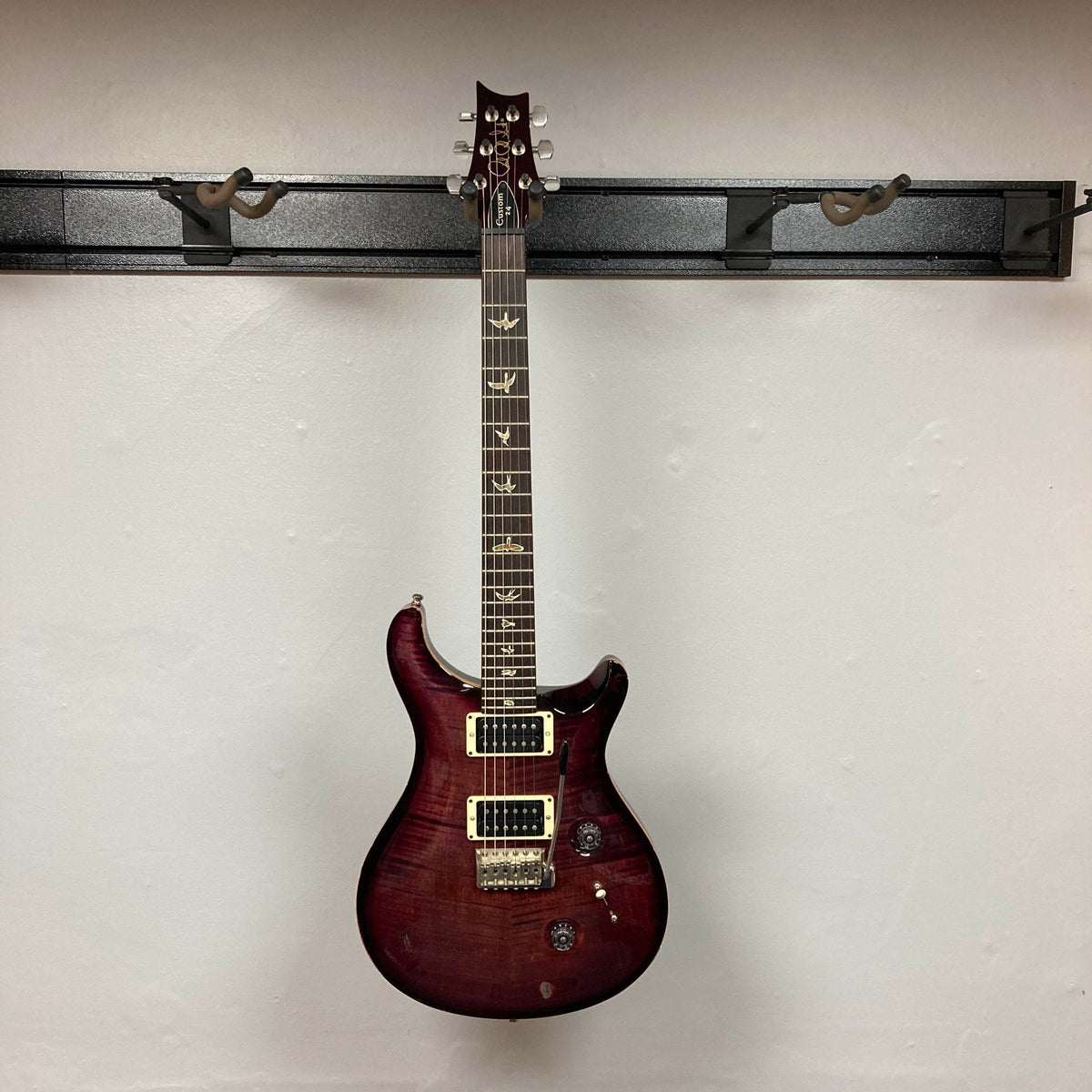 A PRS Custom 24 Angry Larry 2011 electric guitar by PRS, featuring bird inlays on the fretboard and humbuckers, is hanging on a wall-mounted rack against a plain white wall.