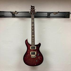 A PRS Custom 24 Angry Larry 2011 electric guitar by PRS, featuring bird inlays on the fretboard and humbuckers, is hanging on a wall-mounted rack against a plain white wall.