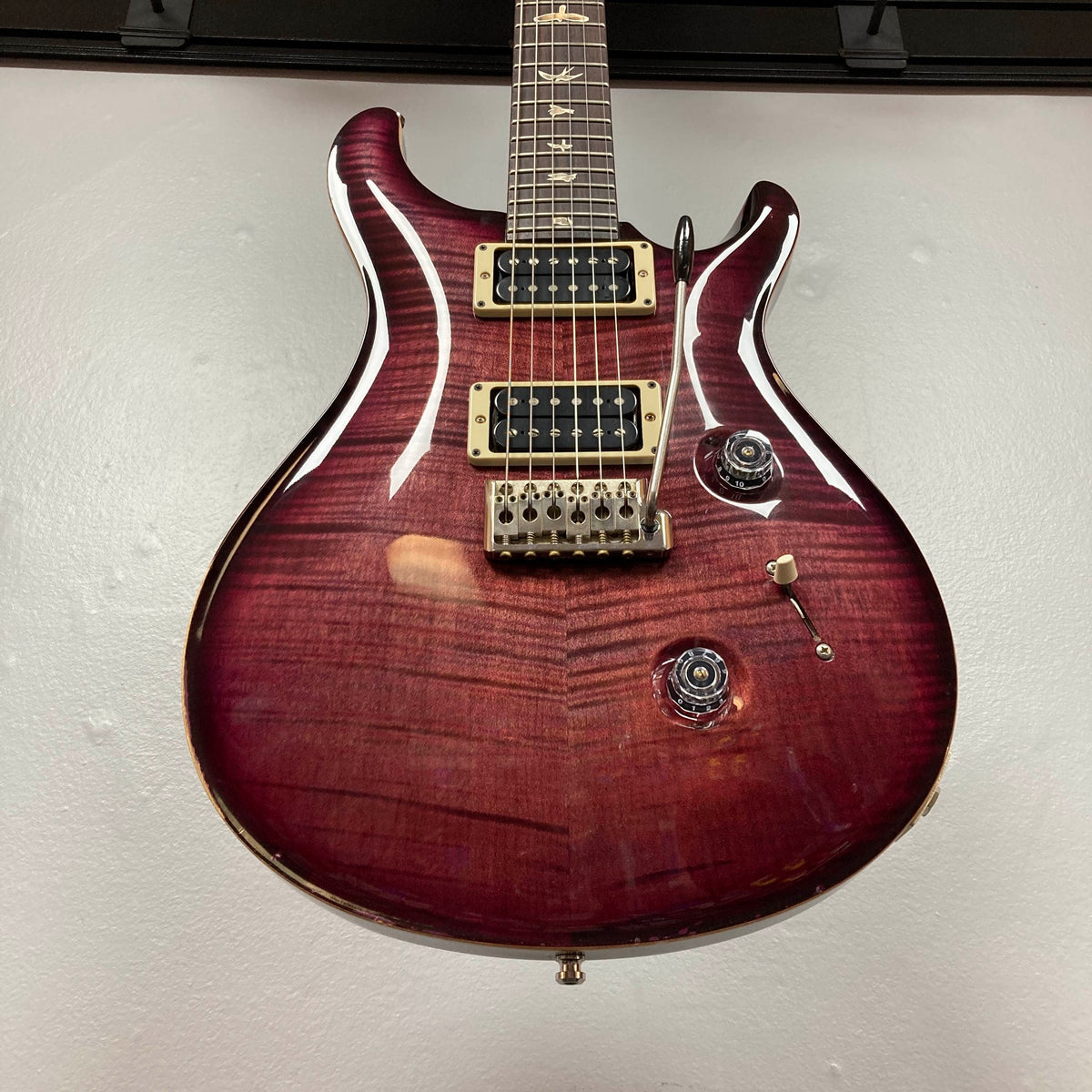 A PRS Custom 24 Angry Larry 2011 electric guitar by PRS, featuring a maroon finish with a glossy flame maple top, two humbucker pickups, a tremolo bridge, and two control knobs, hanging on a wall.