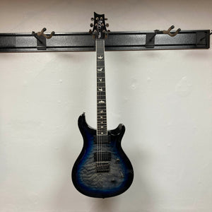 A PRS SE Mark Holcomb - Holcomb Blue Burst w/Gigbag Used Consign hangs on a black wall-mounted holder against a white wall.