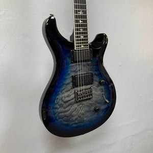 A PRS SE Mark Holcomb - Holcomb Blue Burst w/Gigbag Used Consign electric guitar hangs on a white wall, showing off its stunning finish and bird inlays.