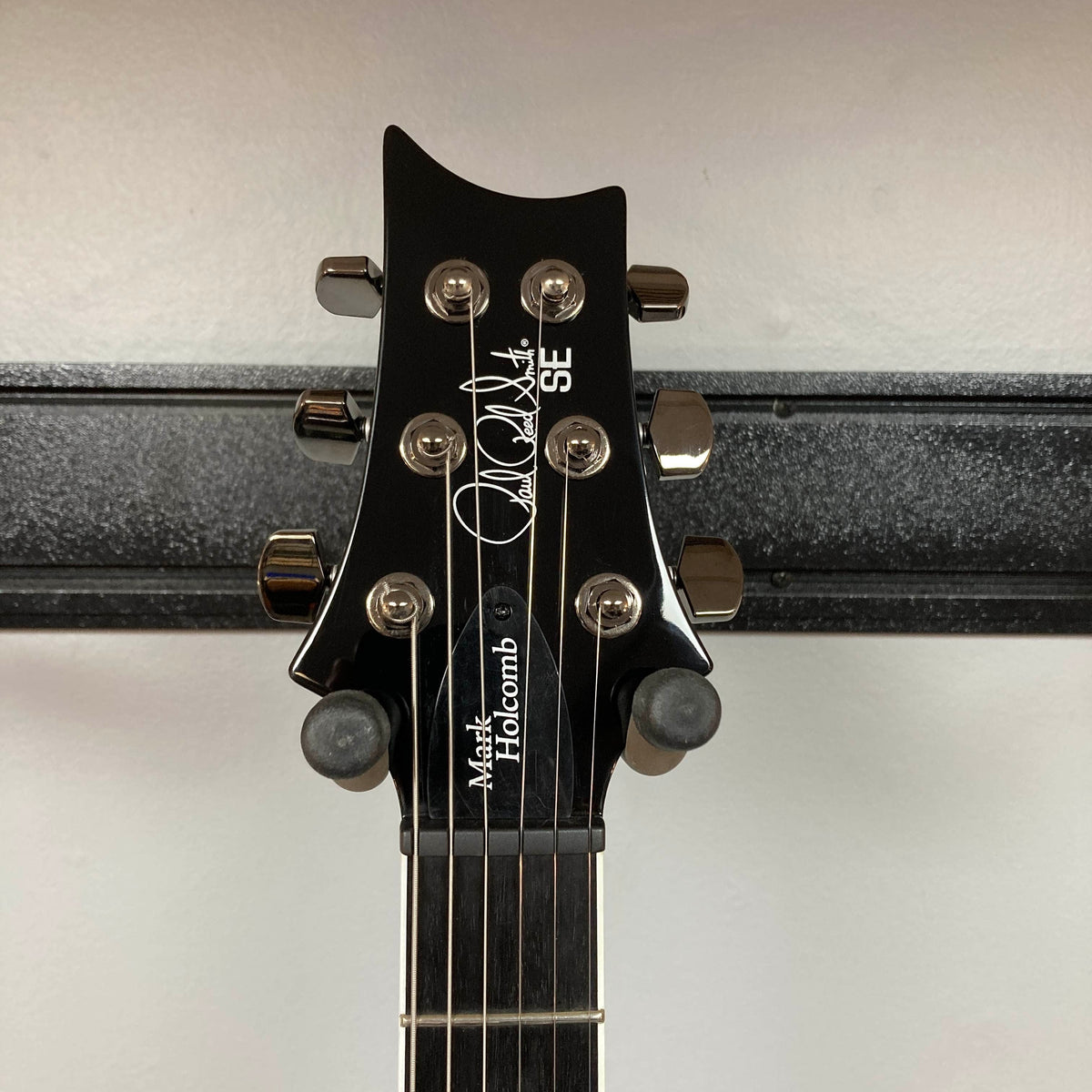 Close-up of the headstock of a PRS SE Mark Holcomb - Holcomb Blue Burst w/Gigbag Used Consign, mounted on a hanger against a white wall.