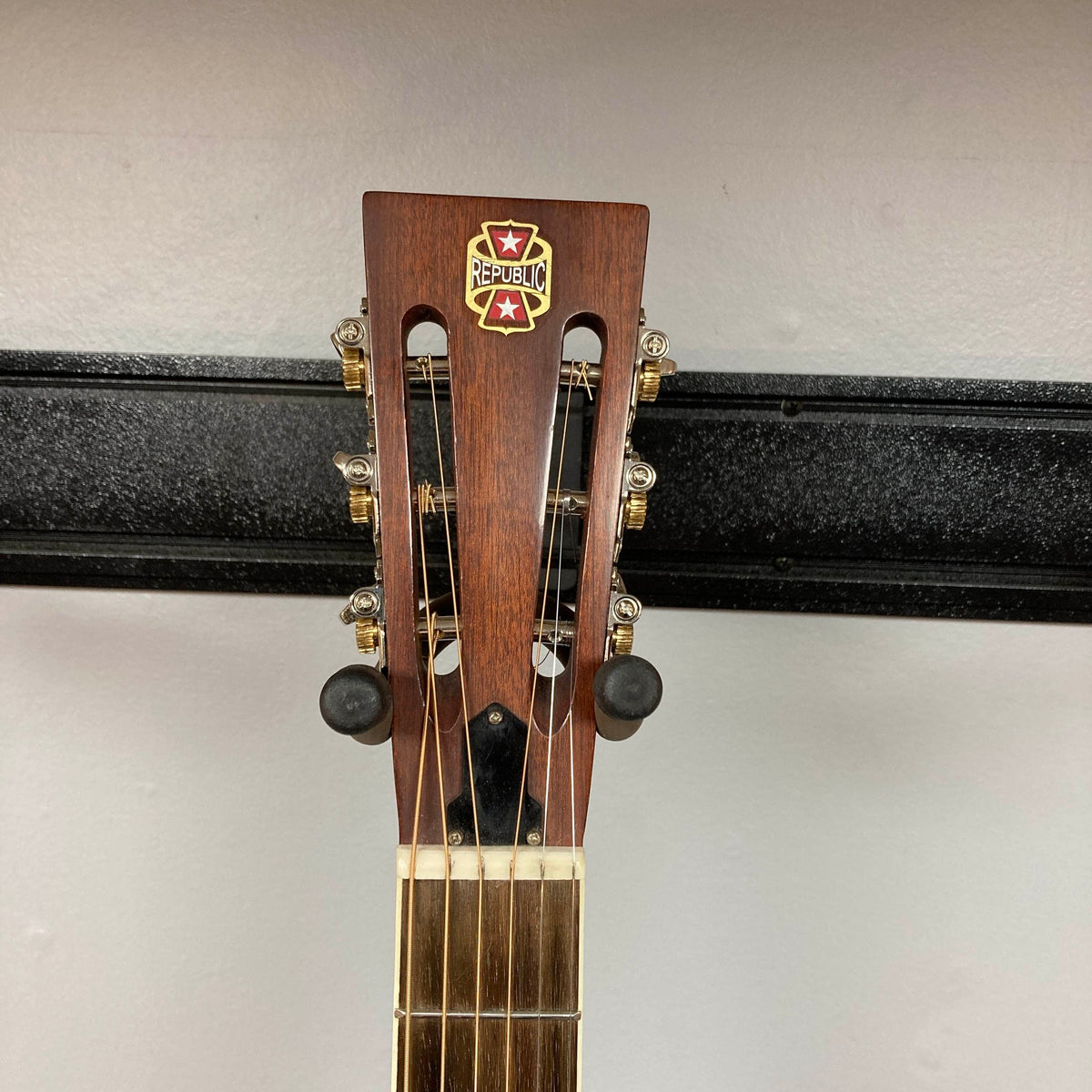Close-up of the Republic Parlor Resolian Antique Bronze Resonator headstock, featuring the Republic logo, six tuning pegs, and a light-colored fretboard. Its single cone design gleams through the antique bronze finish, set against a plain wall and black rack.