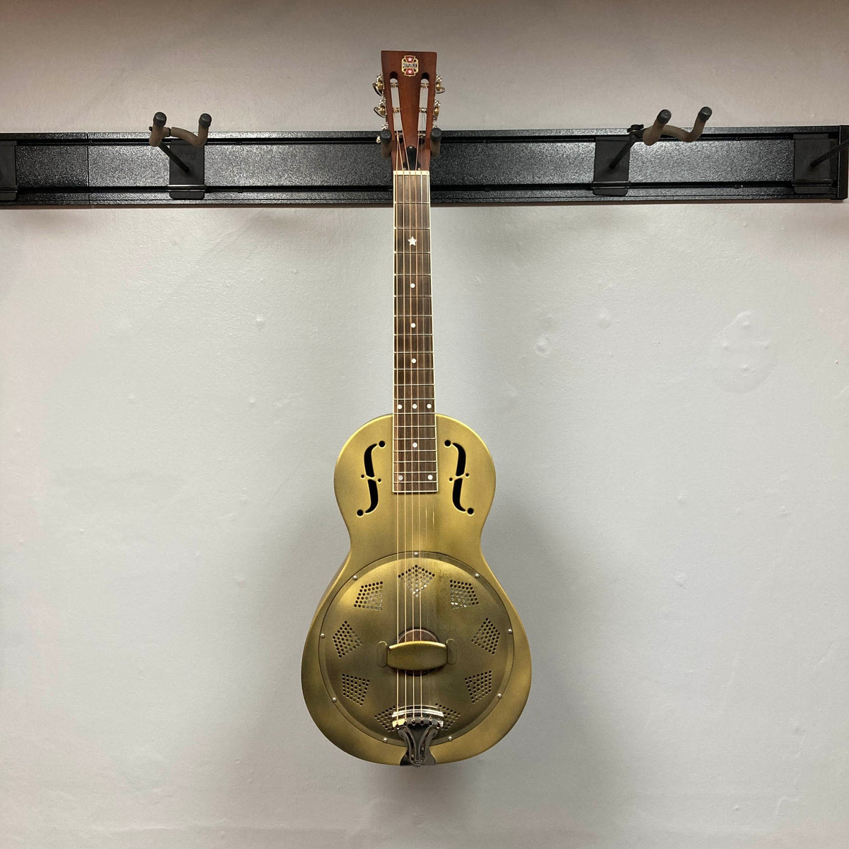 The Republic Parlor Resolian Antique Bronze Resonator w/Case by Republic, featuring a metal body with f-holes and a single cone, hangs on a wall-mounted rack against a white background, showcasing its subtle antique bronze finish.