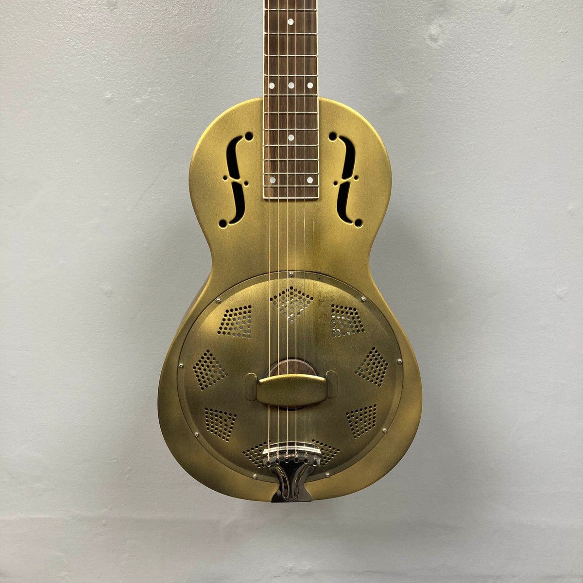 The Republic Parlor Resolian Antique Bronze Resonator w/Case, a brass-bodied six-string guitar featuring f-holes, a single cone, and a metal cover plate, hangs against a plain light gray wall.