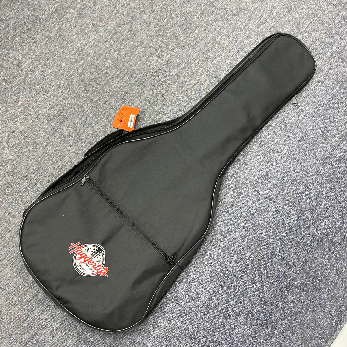 A black soft gigbag with a front zippered pocket and a "Haggleco" logo, perfect for your Reverend Greg Koch Signature Gristle 90 3-Tone Burst electric guitar, rests on a gray carpeted floor.