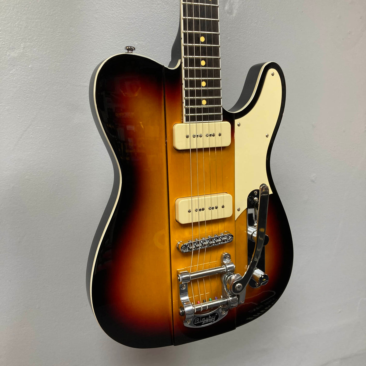 A Reverend Greg Koch Signature Gristle 90 electric guitar in 3-Tone Burst with a white pickguard, two cream-colored Fishman Gristle 90 pickups, Bigsby tremolo, and chrome hardware is shown against a plain gray wall. Brand: Reverend.