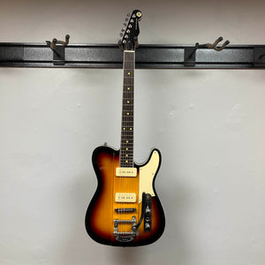 A Reverend Greg Koch Signature Gristle 90 3-Tone Burst electric guitar with white pickguard and metal tremolo arm hangs on a black wall-mounted hook against a plain white wall.