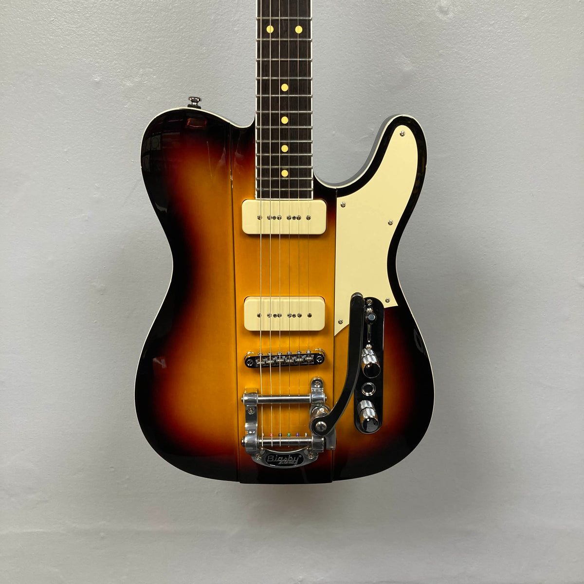 The Reverend Greg Koch Signature Gristle 90 3-Tone Burst electric guitar features two cream Fishman pickups, a white pickguard, and a Bigsby vibrato tailpiece, shown here against a plain gray wall.