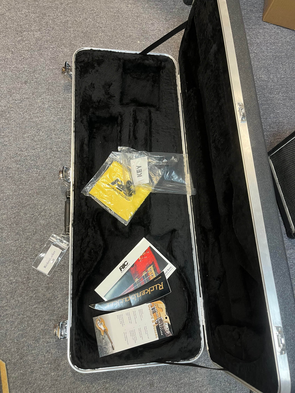A near-mint Rickenbacker 620 Jetglo with its original black velvet-lined case sits on a gray carpet, including a yellow cleaning cloth, key in plastic bag, manuals, and paperwork.