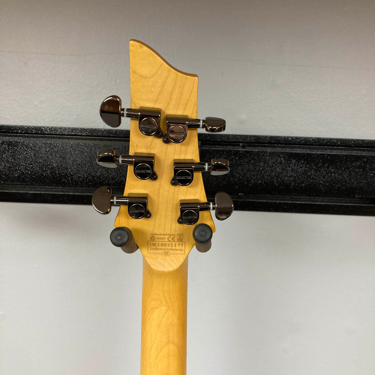 Close-up of the back of a Schecter Extreme Banshee 6 FR CHARCOAL BURST Electric Guitar headstock, featuring six tuning pegs and part of the neck, set against a plain wall. Includes case. Brand: Schecter.