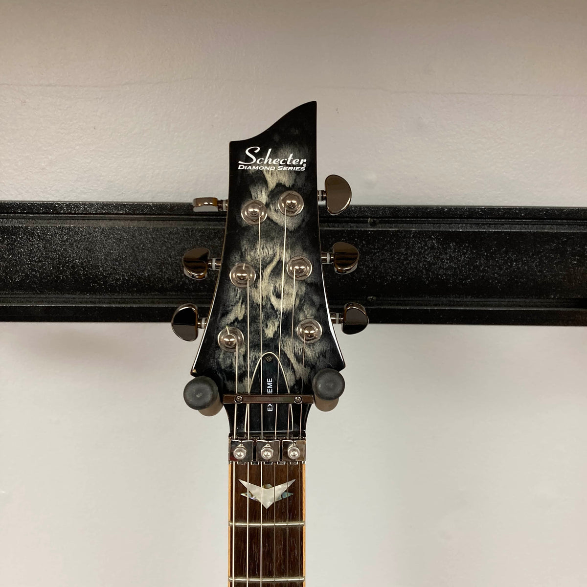 The headstock of the Schecter Extreme Banshee 6 FR CHARCOAL BURST Electric Guitar w/case, featuring six tuning pegs, is shown on a wall rack against a plain background.