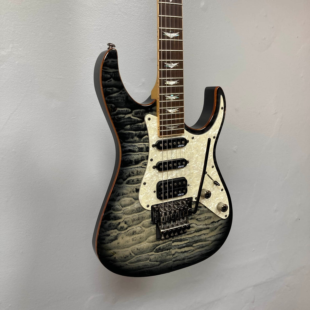 Schecter Extreme Banshee 6 FR CHARCOAL BURST Electric Guitar w/case, featuring a quilted maple top, three pickups, pearloid pickguard, and double-cutaway body, hangs on a light-colored wall.