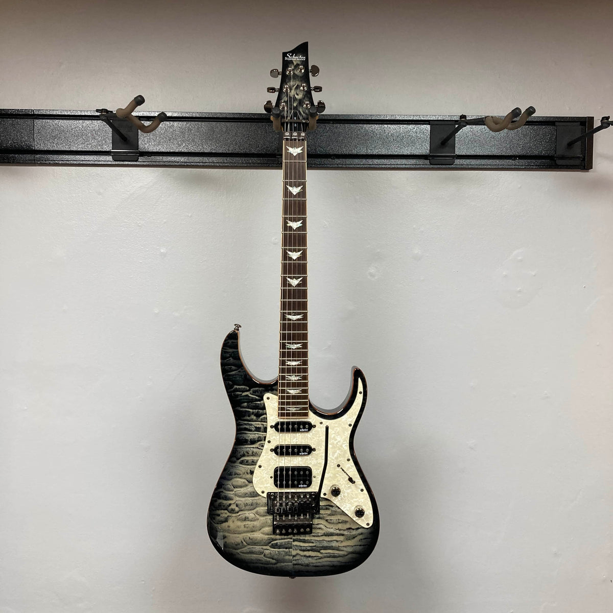 The Schecter Extreme Banshee 6 FR CHARCOAL BURST Electric Guitar w/case, featuring a striking quilted maple finish and bold design, hangs on a wall-mounted rack against a plain white background.