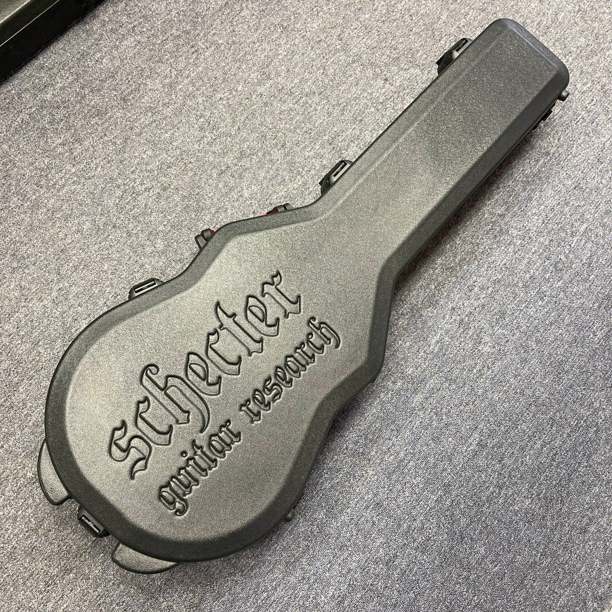 A Schecter Solo-II Custom Trans Black Burst with case, branded "Schecter," lies on a gray carpeted floor. The case is hard, black, and embossed with "Schecter guitar research," made to fit the Solo-II Custom model.
