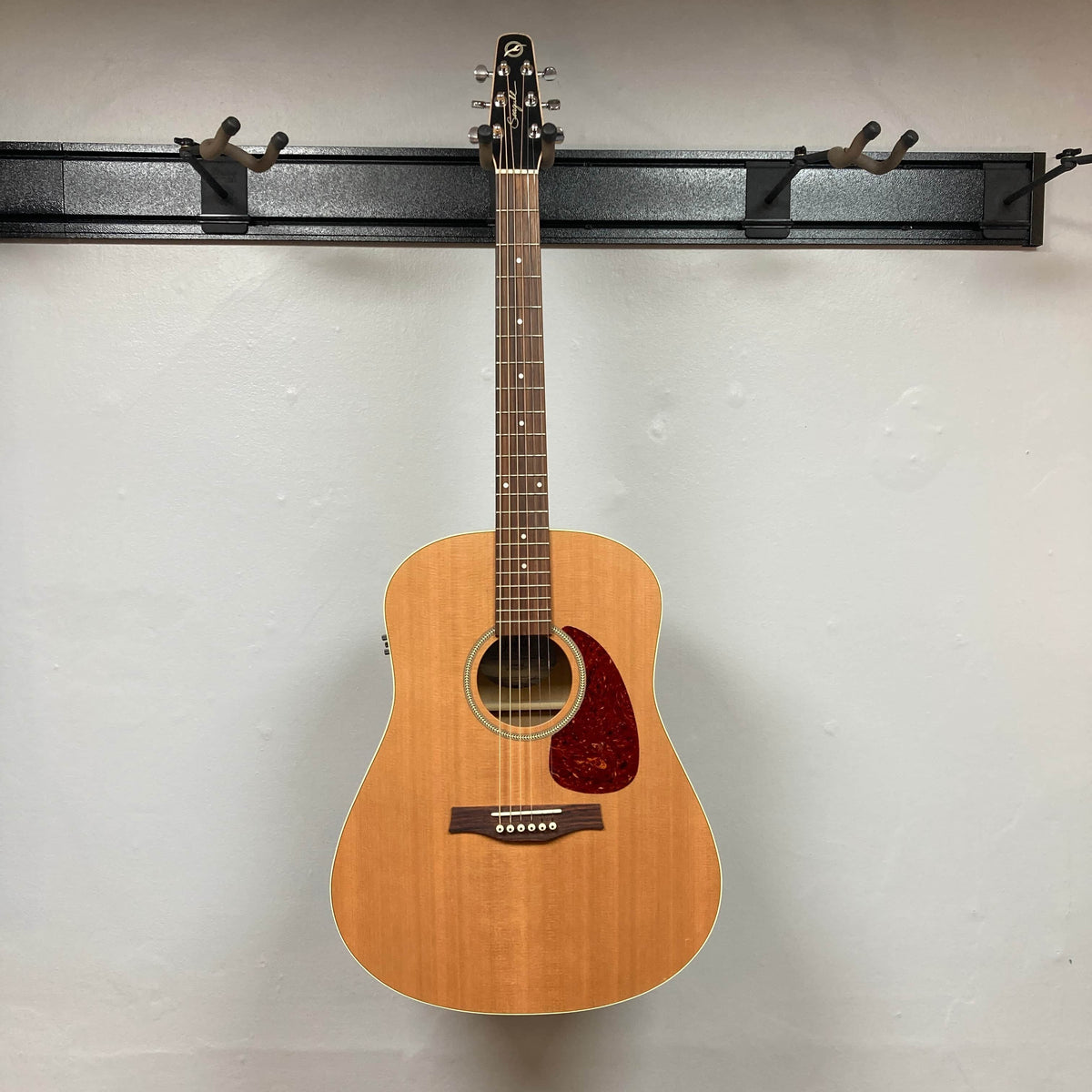 A Seagull S6 QI Slim Acoustic Guitar w/Hard Case (USED/consign) hangs on a wall-mounted rack against a plain white background, available now for consignment sale.