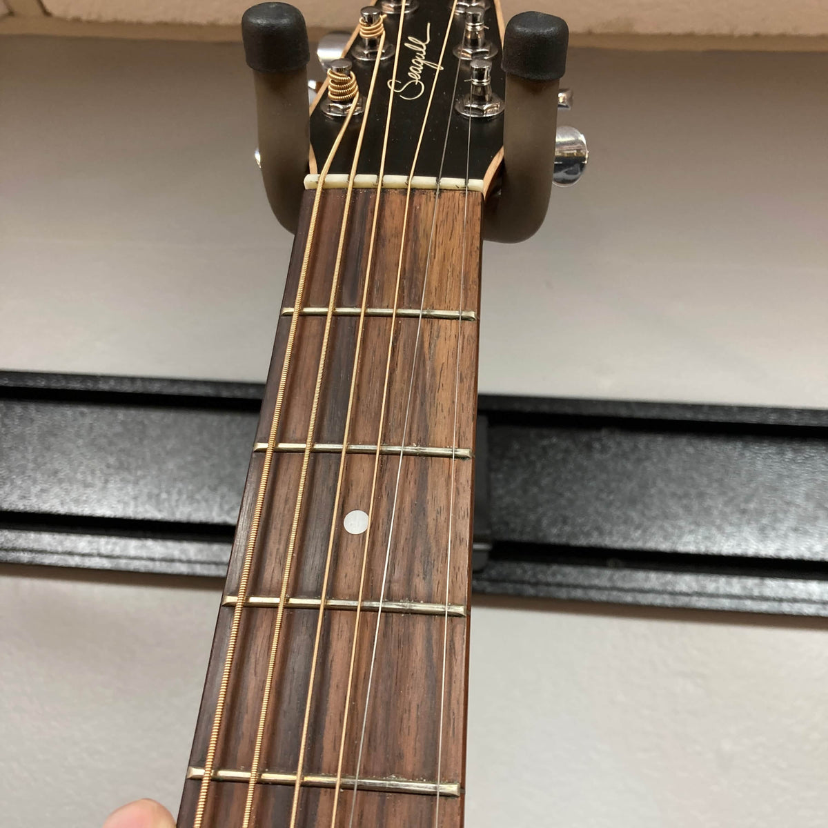 Close-up of a Seagull S6 QI Slim Acoustic Guitar neck with six strings and signature logo on the headstock, displayed on a wall hanger. Includes hard case—ideal for anyone wanting a quality, travel-friendly acoustic guitar.