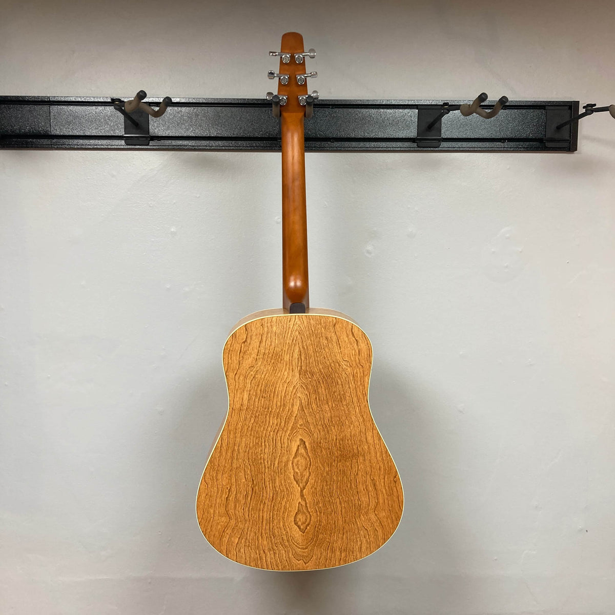 A Seagull S6 QI Slim Acoustic Guitar w/Hard Case - USED/consign hangs by its neck on a black wall-mounted rack, seen from the back against a plain light-colored wall.