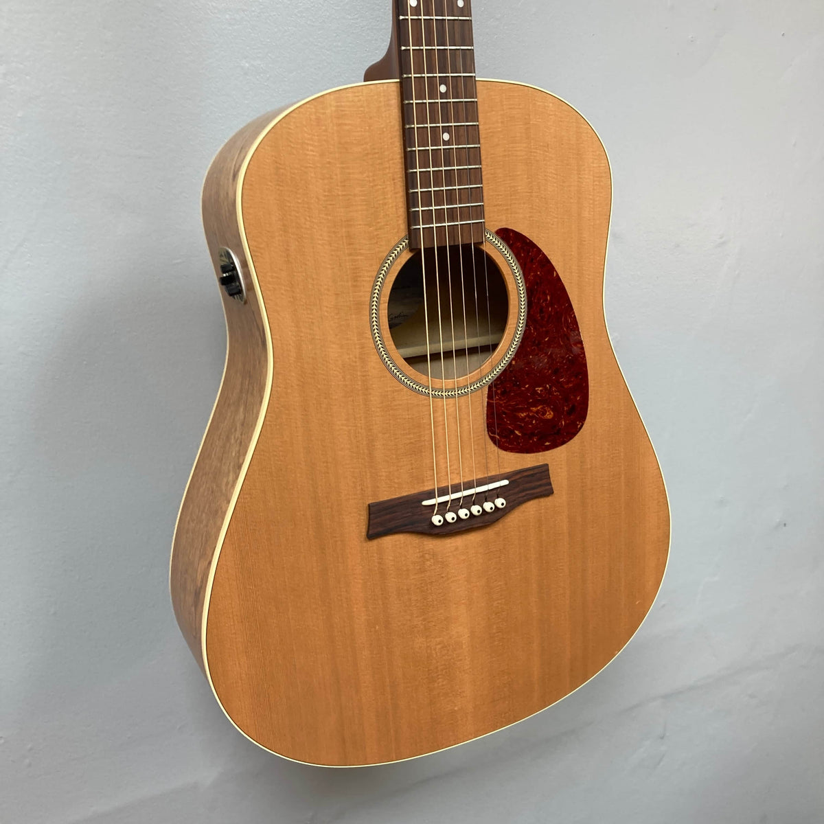 The Seagull S6 QI Slim Acoustic Guitar w/Hard Case - USED/consign features a solid spruce top, six strings, and a reddish-brown pickguard, shown mounted against a light gray wall.