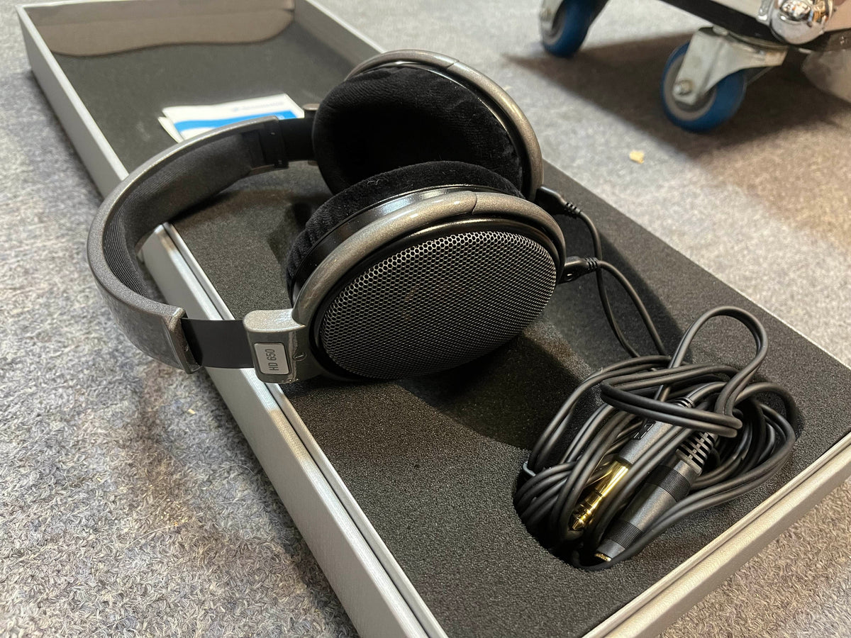 A pair of preowned Sennheiser HD 650 Reference Class headphones, featuring an open-back black design with a coiled cable and adapter, are displayed in a foam-lined case on a grey carpeted surface.
