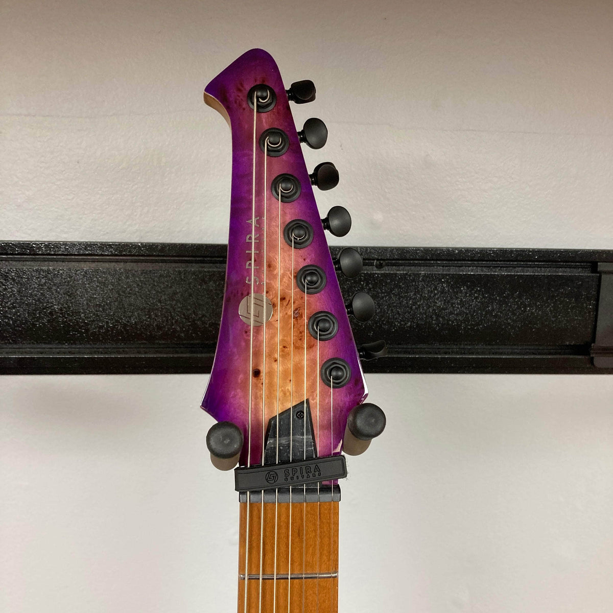 Spira S-457 TPP Trans Purple Burst Electric Guitar headstock, featuring black tuning machines and Spira logo, mounted on a wall.