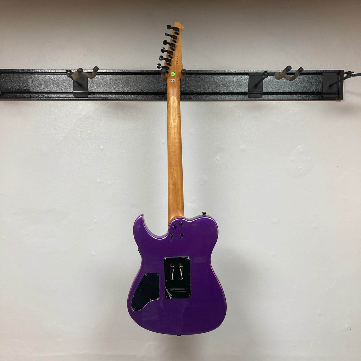 A Spira T-420 PP electric guitar by Spira, in purple with a roasted maple neck and missing backplates that show the wiring, hangs on a wall-mounted rack and is photographed from the back against a white wall.