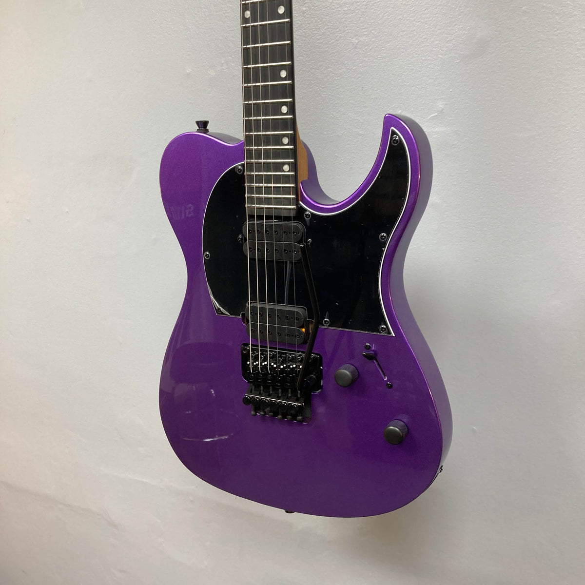 A Spira T-420 PP electric guitar in purple, featuring high-output humbuckers, black pickguard and hardware, and two control knobs, hangs against a plain white wall.