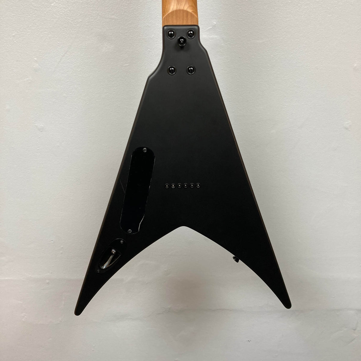 The image shows the back of a Spira V-400 MBK electric guitar by Spira, featuring a black V-shaped body and natural wood neck against a light wall, celebrated for its tuning stability.