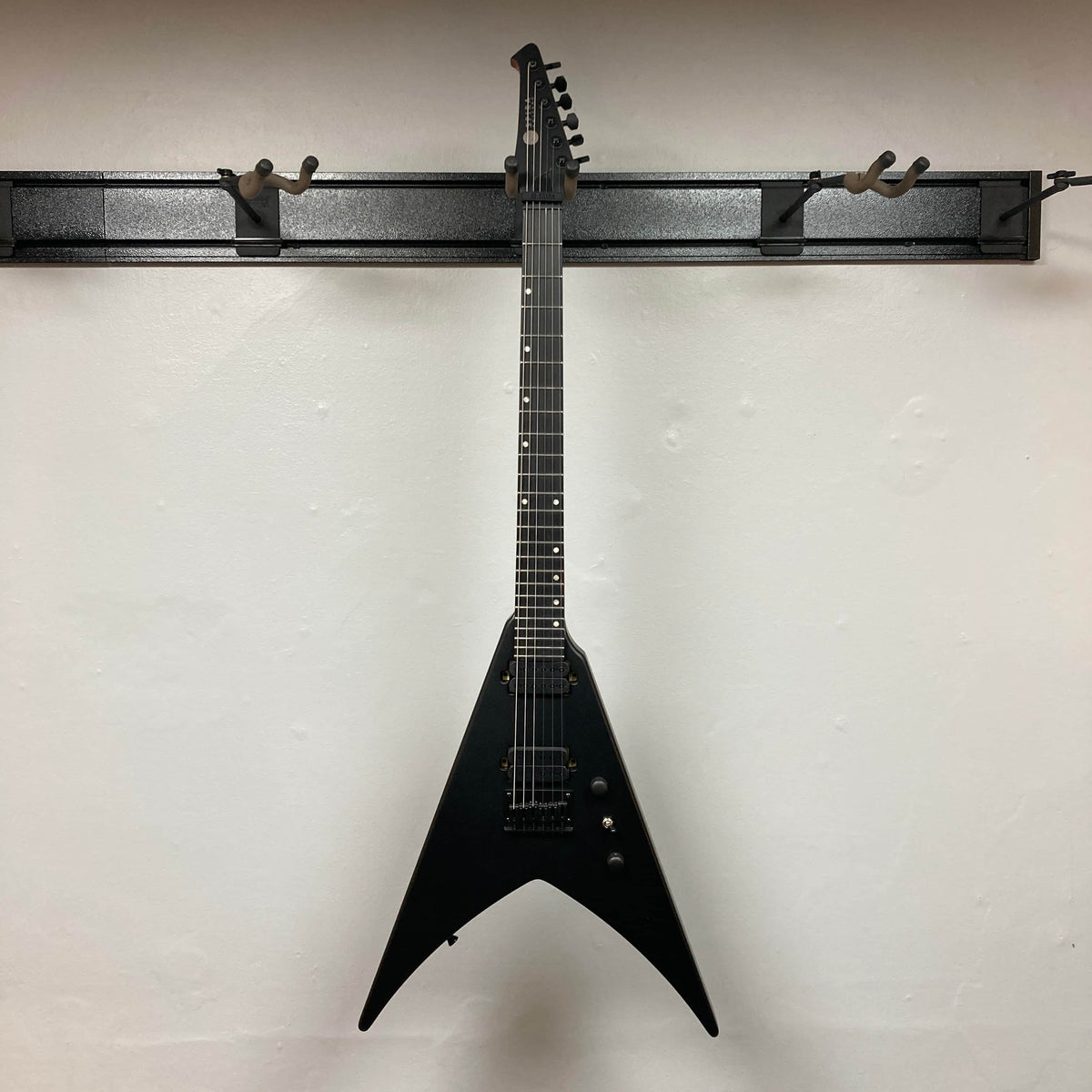 A Spira V-400 MBK electric guitar by Spira, featuring high-output humbuckers and a V-shaped black body, hangs on a wall-mounted rack against a plain white background.