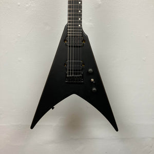 A Spira V-400 MBK electric guitar with a matte black V-shaped body, high-output humbuckers, three knobs, and a switch is photographed against a plain white wall.