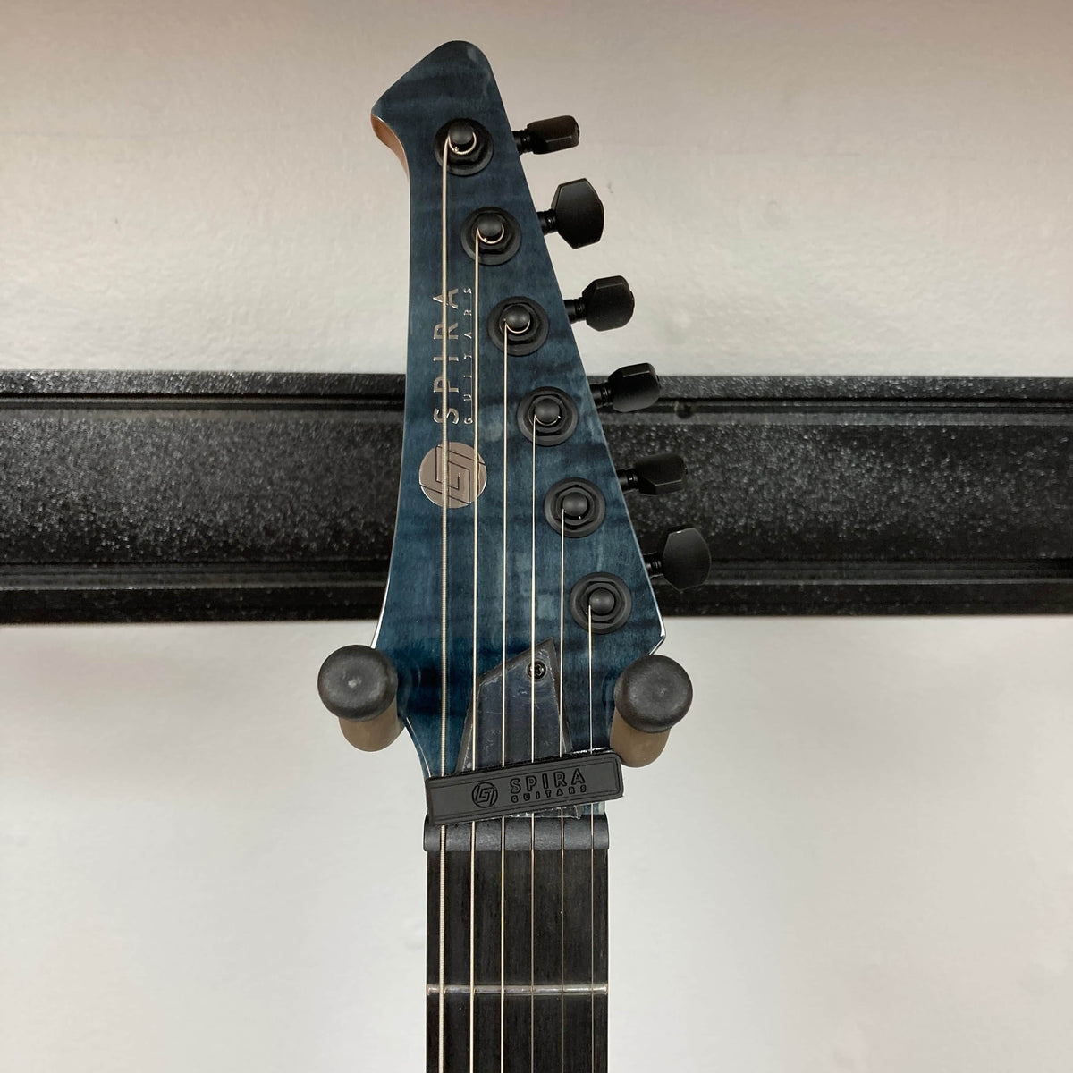 Close-up of a blue Spira X-500 TBK E electric guitar headstock with six locking tuners, set against a white wall and black guitar hanger.