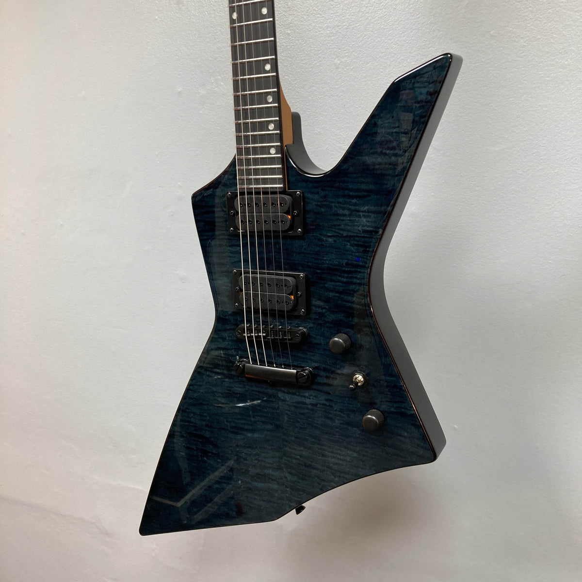The Spira X-500 TBK E by Spira features a dark blue, angular, asymmetrical body with high-output humbuckers, black hardware, and locking tuners—an ideal metal guitar—shown against a white wall.