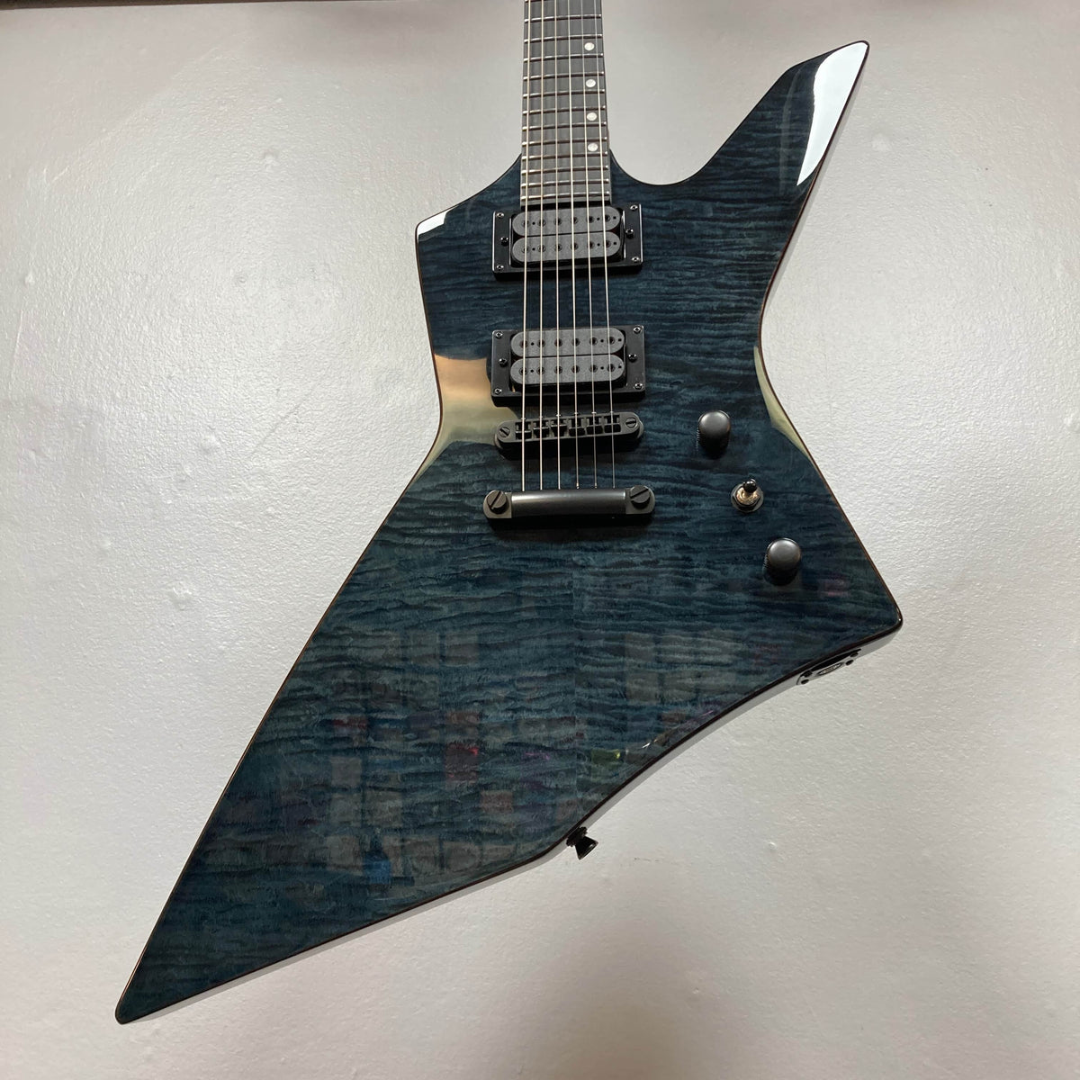 The Spira X-500 TBK E by Spira is an electric metal guitar with a sharp, angular body, dark blue wood grain finish, high-output humbuckers, and three control knobs set against a plain light-colored background.