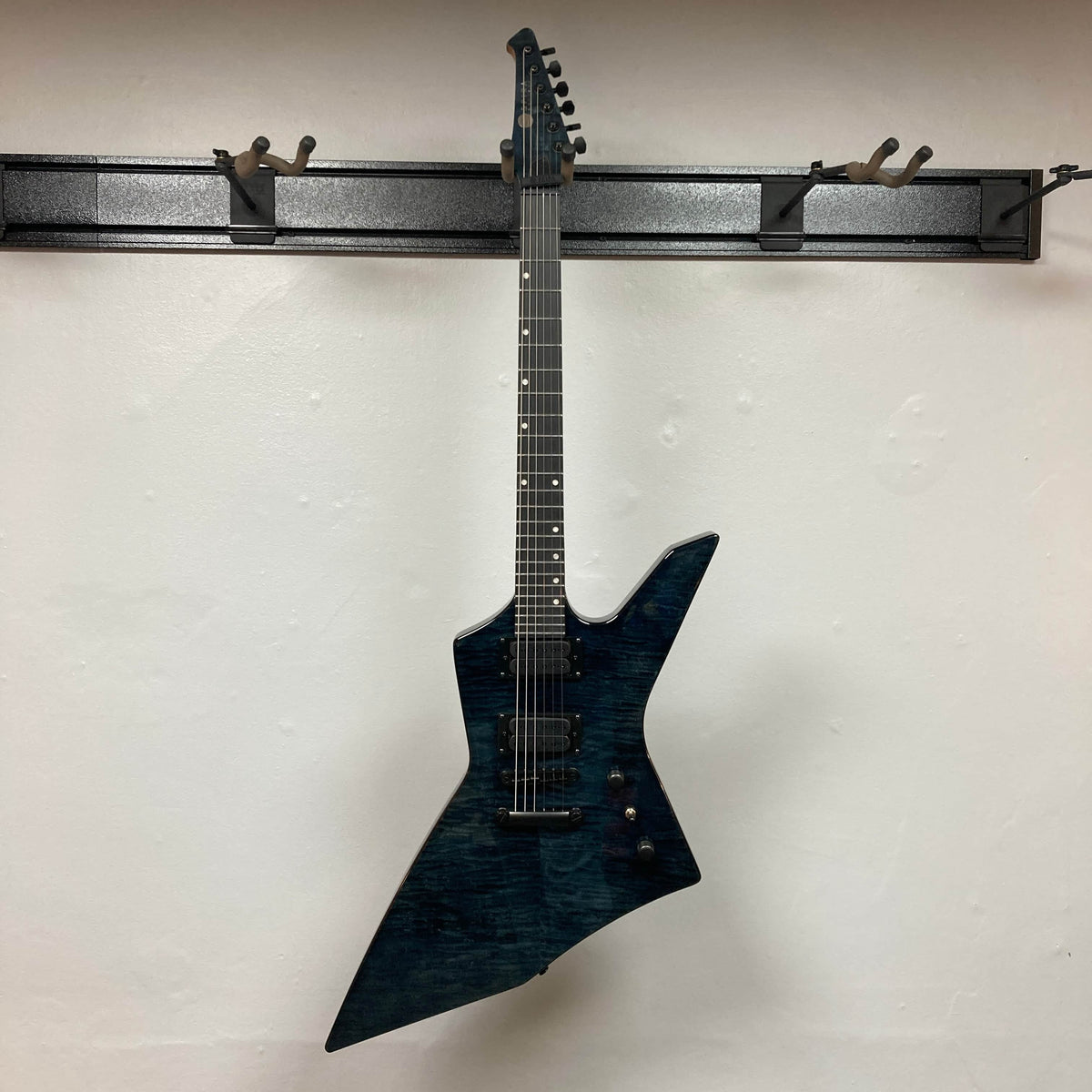 A Spira X-500 TBK E guitar by Spira, featuring a dark blue metal finish, angular asymmetric body, and high-output humbuckers, hangs on a wall-mounted rack against a white background.