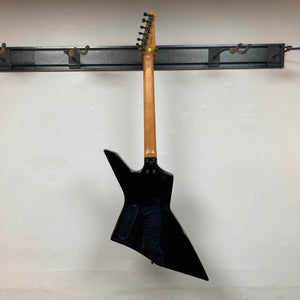 A Spira X-500 TBK E black metal guitar with an angular body and fast neck hangs by its neck on a wall-mounted rack against a plain white wall.