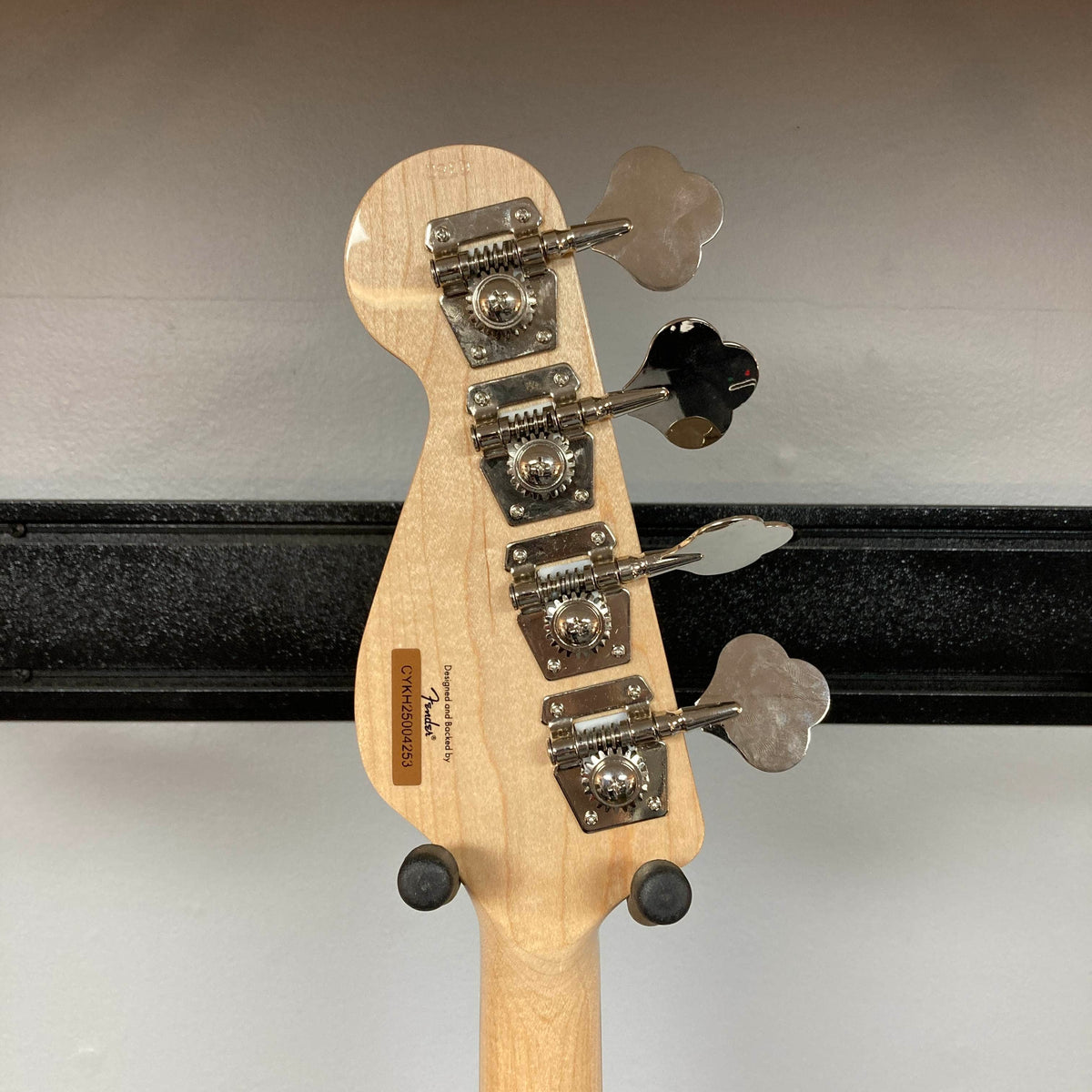 The back of a Squier Paranormal Rascal Bass HH Sherwood Green Refurb headstock, displaying four tuning pegs and a serial number, is mounted on a wall hanger against a white background—highlighting the distinctive short scale bass design from Squier.