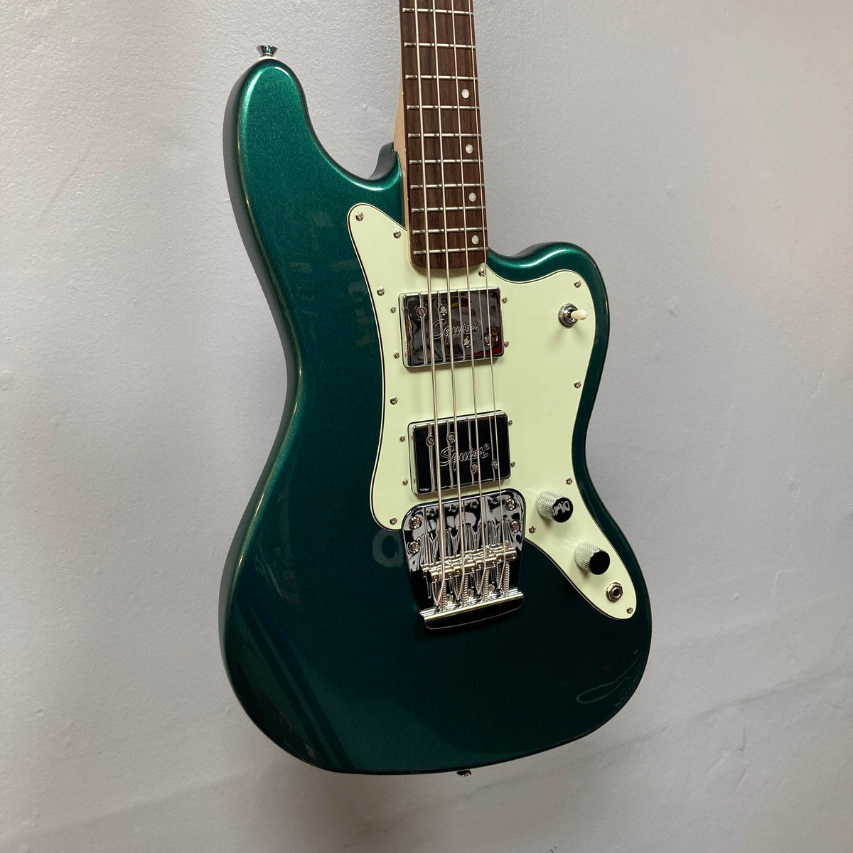A Squier Paranormal Rascal Bass HH in Sherwood Green with a white pickguard, two pickups, and a chrome bridge hangs on a plain light-colored wall.