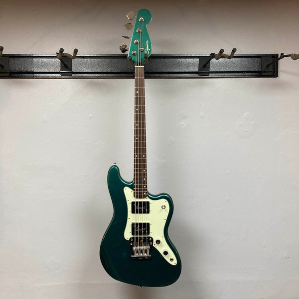 A Squier Paranormal Rascal Bass HH Sherwood Green Refurb, featuring a white pickguard, Fender Designed Wide Range humbuckers, four control knobs, and a chrome bridge, stands against a plain light gray wall.