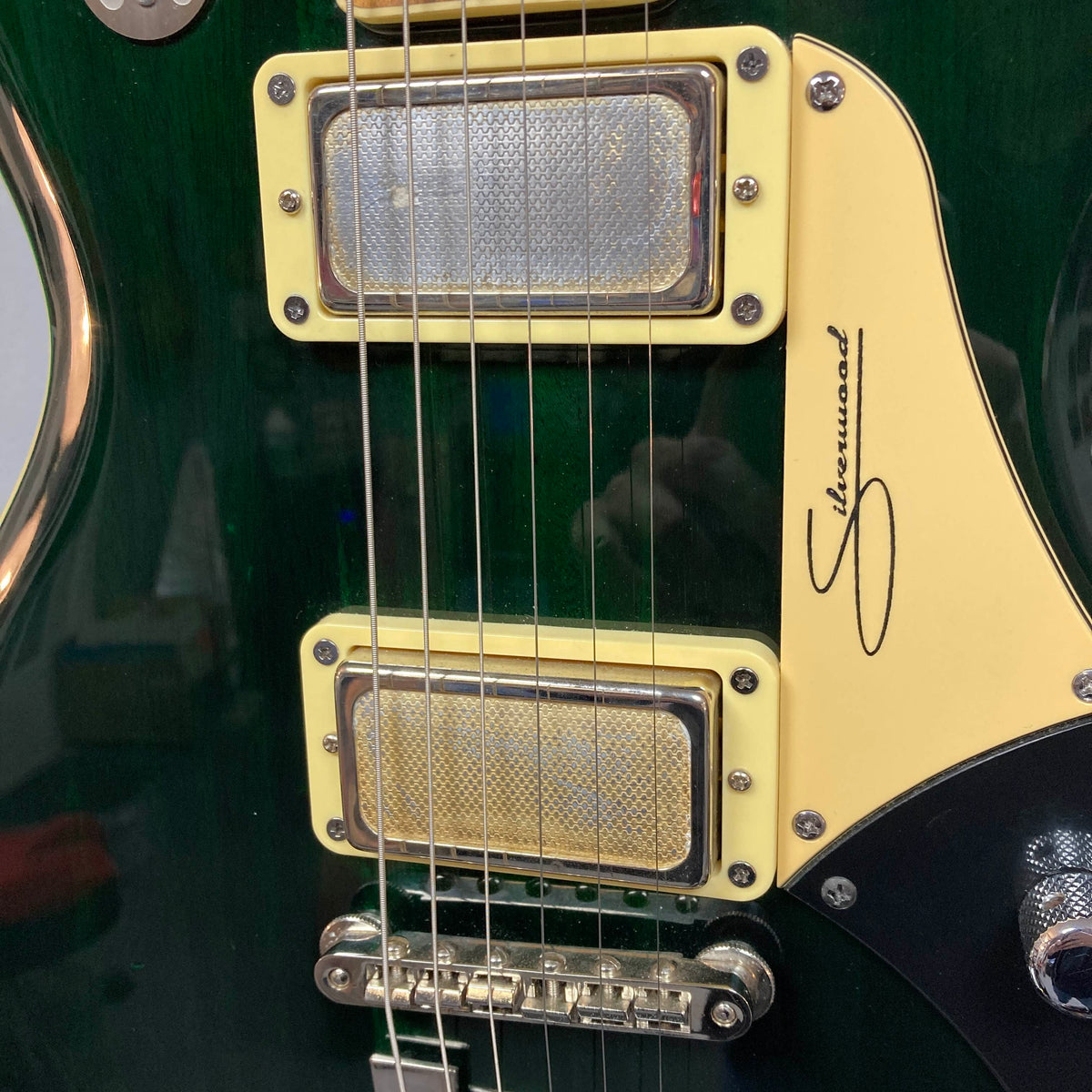 Close-up of a Supro 1296 Silverwood guitar in British Racing Green, featuring two Gold Foil pickups, six strings, a green body, and a cream pickguard adorned with the "Sherwood" signature and stylized 'S' logo.