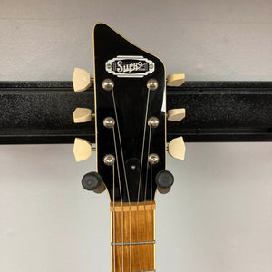 Close-up of the Supro 1296 Silverwood British Racing Green headstock with six tuning pegs, a black faceplate, and wood neck, resting on a guitar hanger against a gray wall.