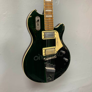A Supro 1296 Silverwood in British Racing Green with gold hardware, cream-colored Gold Foil pickups, and a signature on the body is mounted on a light gray wall.