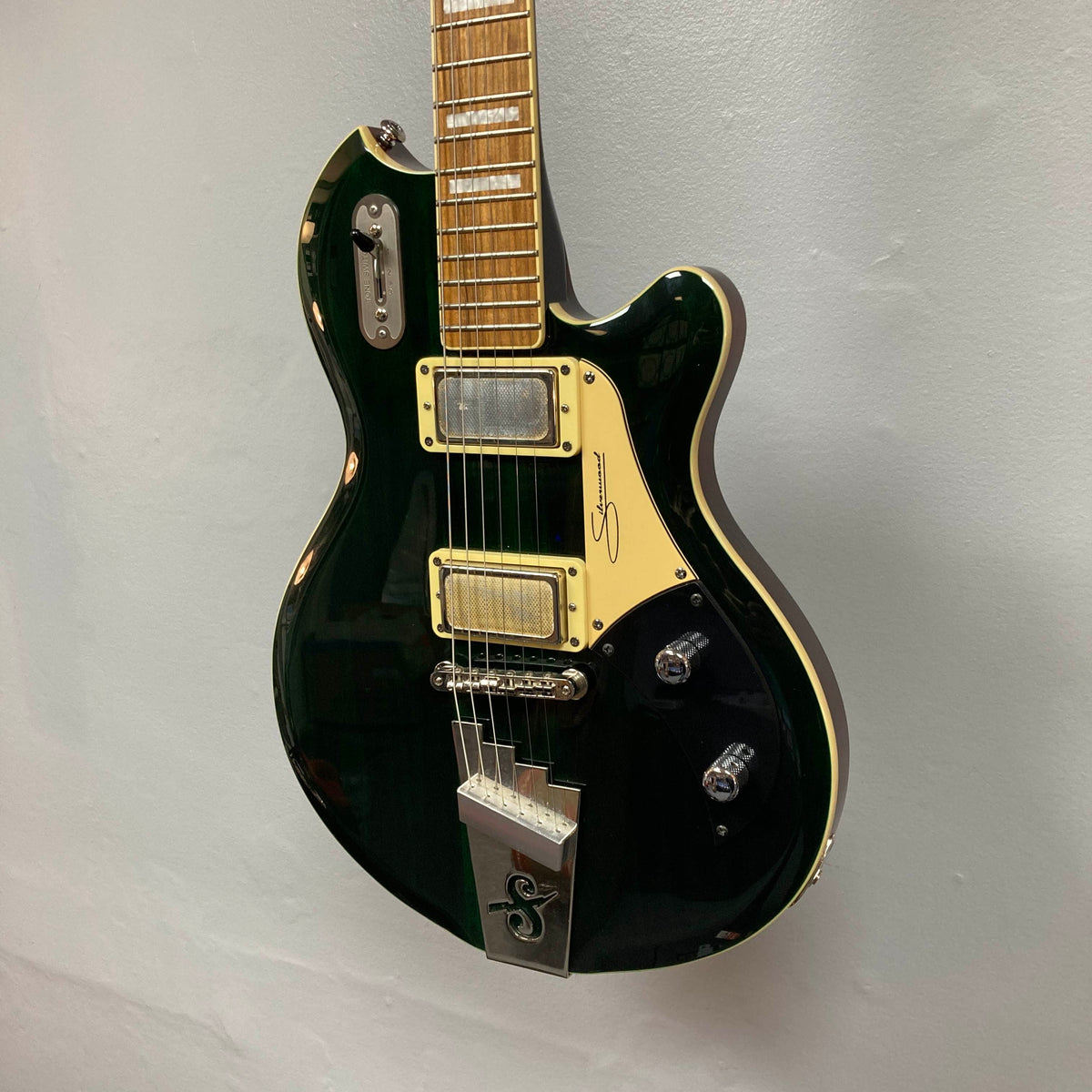 A Supro 1296 Silverwood electric guitar in British Racing Green with a cream pickguard, chrome hardware, distinctive Gold Foil pickups, and control knobs is shown hanging against a plain light gray wall.